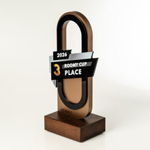 Load image into Gallery viewer, A modern award trophy shaped like a padlock, with a metallic bronze and black body, a dark wood base, and a ribbon label reading '2026 ROOMY CUP PLACE 3' and 'STAGE 1'.