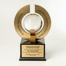 Load image into Gallery viewer, A custom corporate trophy featuring a large brushed gold circular ring with engraved text, a clear glass globe at the center, and a black rectangular base with a gold plaque containing personalized text.